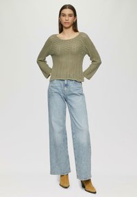 Olijfgroene gebreide trui met een gestructureerd patroon, wijdere mouwen en een relaxte pasvorm, gecombineerd met lichtblauwe high-waisted jeans en tan enkelboots.