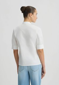 Blouse blanche à manches courtes avec un col montant, présentant une fermeture à boutons visible au dos. Portée avec un pantalon en denim bleu clair.