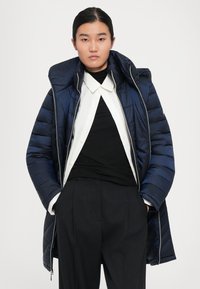 Jongere draagt een marineblauwe pufferjack over een witte blouse en een zwart topje, met zwart gestreepte broek, handen in de zakken.