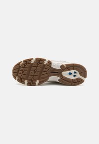 Weiße Laufschuhe mit einer strukturierten Gummisohle und braunen sowie blauen Akzenten. Tiefe Rillen für Traktion und Unterstützung.