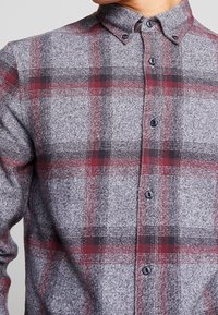 Camicia a quadri con sfondo grigio e quadretti rossi e neri. Il tessuto ha una finitura strutturata ed è dotata di bottoni e colletto.