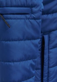 Blå vadderad jacka med texturerad tyg, horisontellt quiltat mönster, en ficka med dragkedja och justerbar elastisk snodd i nederkant.