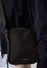 Borsa a tracolla nera Calvin Klein con texture liscia, forma quadrata, tracolla regolabile e un logo discreto nella parte inferiore.