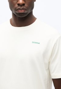 Άνδρας που φοράει ένα απλό λευκό T-shirt με πράσινο κείμενο "30904" στο αριστερό μέρος του στήθους, εμφανιζόμενος από τον λαιμό μέχρι το μέσο του κορμού με λευκό φόντο.