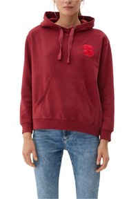 Sweat à capuche rouge en tissu doux, avec une poche avant et un logo brodé texturé sur la poitrine, porté avec un jean bleu.