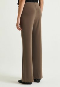 Pantaloni marroni a gamba larga in tessuto liscio, con vita alta, dettagli sulle cuciture laterali e calzature nere posizionate in basso.