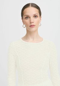 Haut en dentelle crème à manches longues avec un motif délicat, un col rond et un tissu texturé. Design élégant avec des détails subtils.