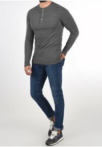 Man in een donkergrijze lange mouwen henley-shirt, blauwe opgerolde jeans en zwart-witte Nike sneakers, staande met één hand in zijn zak.