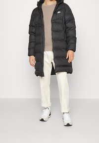 Svart puffjacka med dragkedja fram, stora fickor och huva, kombinerad med en beige tröja och vita byxor, bärs med sneakers.