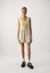 Svetlo béžový pletený top bez rukávov s vlnitými okrajmi, kombinovaný so svetlými šortkami a hnedými platformovými topánkami. Modelka stojí na neutrálnom pozadí.