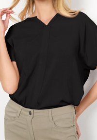 Donna che indossa una blusa nera a maniche corte con scollatura a V infilata in pantaloni beige, con una mano che tocca i capelli e l'altra posata sull'anca.