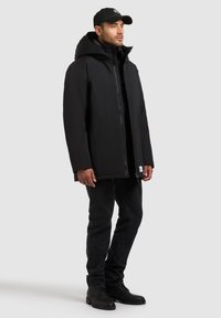 Svart isolerad jacka med hög krage och huva, framzipper och avslappnad passform. Bar över en svart skjorta och ihop med svarta jeans och stövlar.