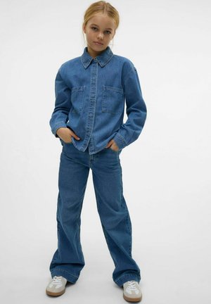 Chemise en jean bleu clair avec un col, deux poches frontales, manches longues. Associée à un jean bleu à jambe large et des baskets blanches.