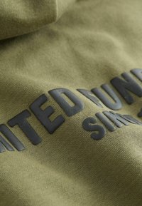 Közelkép oliva zöld szöveten, amelyen fekete kiemelt szöveg olvasható, a "UNITED FUND SIAM" részlete érzékelhető a textúrázott felületen.