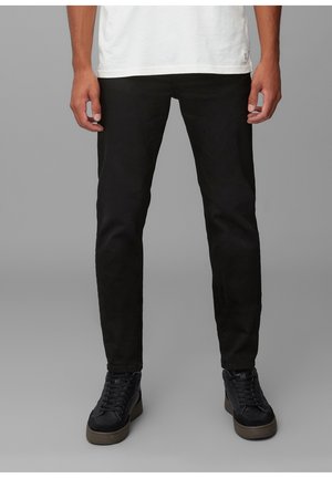 Homme portant un pantalon noir slim, des baskets noires avec des semelles marron et un t-shirt blanc, debout devant un fond gris uni.