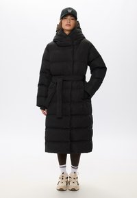 Giacca a vento nera con vita cintata, cappuccio oversize e texture trapuntata. Indossata con pantaloni marroni e sneakers beige.