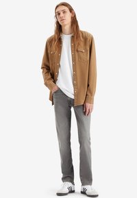 Brun knappet skjorte over en hvit t-skjorte, satt sammen med grå jeans og svart-hvite joggesko. Casual stil med to brystlommer.