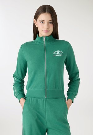 Giovane donna con lunghi capelli castani che indossa una felpa verde con zip e pantaloni coordinati con la scritta "Louisville Kentucky" sul petto.