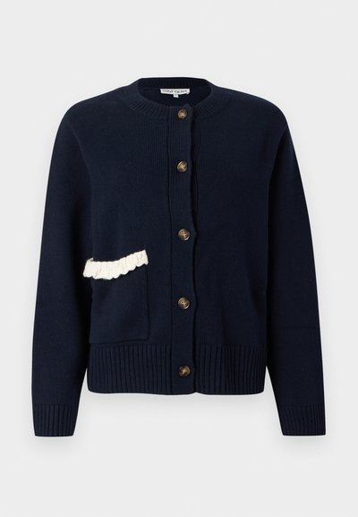 Πλεκτή ζακέτα σε ναυτικό μπλε με στρογγυλή λαιμόκοψη, κουμπιά μπροστά, rib μανσέτες και τελείωμα, και μια λευκή βολάν λεπτομέρεια στην αριστερή τσέπη.