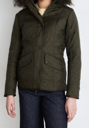 Femme portant une veste matelassée vert foncé avec des poches à boutons-pression et un jean bleu foncé sur un fond uni.