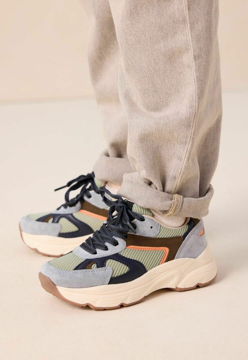 Persoon in beige broek en multicolore sneakers met zwarte veters, met grijze, groene, bruine en oranje accenten op een lichte vloer.