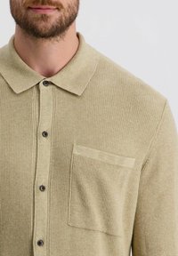 Helles beiges Strickpolohemd mit strukturiertem Muster, Knopfverschluss, Kragen und Brusttasche. Verfügt über dunkle Knöpfe und eine lockere Passform.
