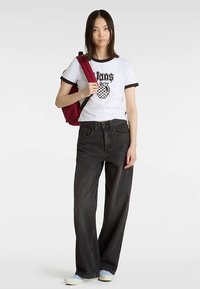 T-shirt blanc avec accents noirs et motif ananas, associé à un jean noir taille haute à jambes larges et des chaussures légères bleu clair. Sac à dos rouge.