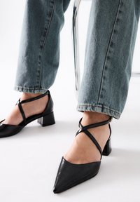 Zapatos de charol negros con punta afilada, tacón bajo y tiras cruzadas en el tobillo, combinados con unos jeans cortos de color azul claro.