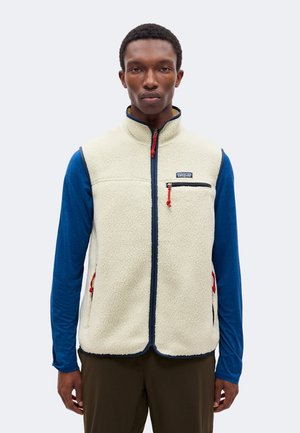 Un homme porte un gilet polaire Patagonia couleur crème, bordé de bleu et doté de tirettes de fermeture éclair rouges, sur une chemise bleue à manches longues.