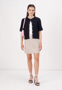 Marineblauwe gebreide cardigan met structuur, korte mouwen, knoopsluiting, gecombineerd met een lichtbeige geribde rok en zwarte hakken met stippenaccenten.