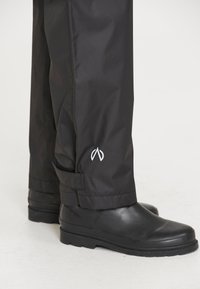 Vandtætte sorte bukser lavet af glat stof, slim fit, med et hvidt logo nær kanten og justerbare stropper. Båret med sorte støvler.