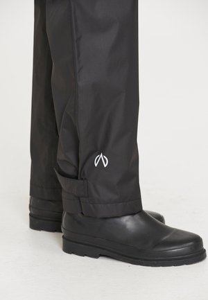 Vandtætte sorte bukser lavet af glat stof, slim fit, med et hvidt logo nær kanten og justerbare stropper. Båret med sorte støvler.