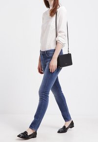 Blusa bianca a maniche lunghe con dettagli a volant, abbinata a jeans blu a taglio slim e mocassini neri in vernice. Una borsa a tracolla nera completa l'outfit.