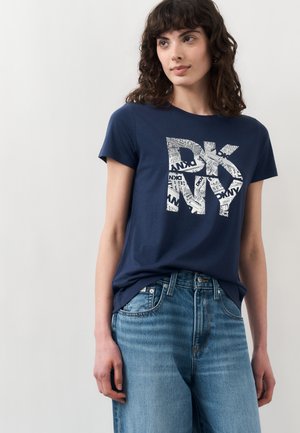 Kvinde iført mørkeblå DKNY T-shirt med stort hvidt logo og lyseblå jeans, stående mod en ensartet lysegrå baggrund.