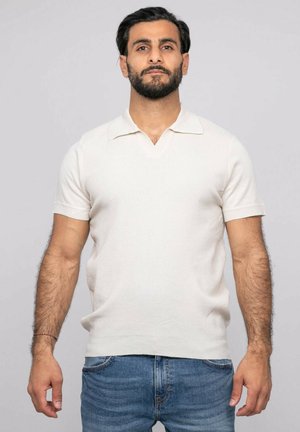 Homme barbu portant un polo blanc en maille à manches courtes et un jean bleu, debout devant un fond clair uni.