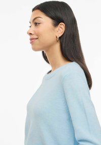 Femme aux longs cheveux bruns portant des boucles d'oreilles créoles dorées et un pull bleu clair, souriant doucement, montrée de profil sur un fond blanc.