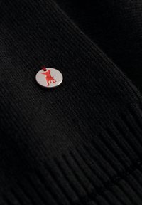 Zwarte gebreide stof met een zilveren ronde metalen speld met een rode silhouetontwerp. Speld bevestigd met een dunne rode draad.