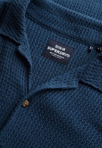 Mørkeblå tekstureret strik polo skjorte med krave, der har en enkelt knaplukning og et label med teksten "SUPERDRY BRITISH DESIGN."