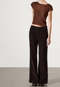 Ruskea metallinhohtoinen lyhythihainen paita, jossa on teksturoitu neulekuvio, yhdistetty tummanruskeisiin leveälahkeisiin corduroyhousuihin. Molemmat vaatteet ovat istuvia ja rentoja.