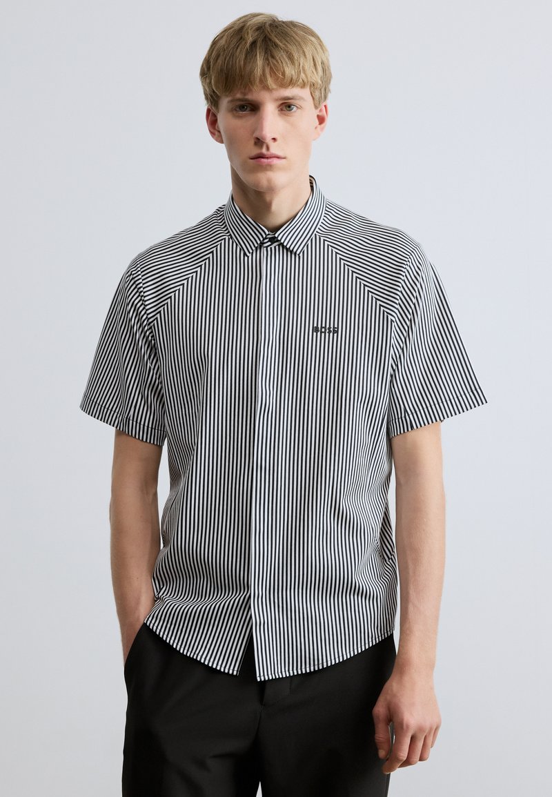 Cămășuță cu mâneci scurte în dungi verticale alb-negru, având un guler cu nasturi și un mic logo pe partea stângă a pieptului.