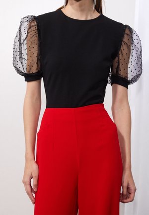 Haut noir à manches courtes bouffantes en mesh transparent à pois noirs, associé à un pantalon rouge taille haute. Texture de tissu lisse.