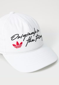 Balta kepurė su juodu išsiuvinėtu tekstu "Original in the Ring" ir raudonu Adidas logotipu. Lygus audinys, suapvalintas kraštas, šešių panelių dizainas.