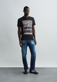 Maglietta nera in cotone con testo grafico bianco e rosa; jeans di denim blu con look sbiadito e dettaglio del logo; sneakers nere con strisce bianche.