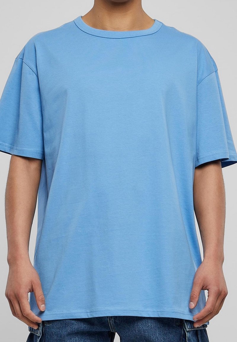 Homme portant un t-shirt à manches courtes bleu ciel ample et un jean en denim bleu foncé, se tenant devant un fond uni.