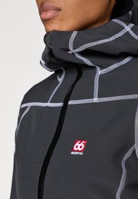 Jacket cinza com fecho de correr e gola alta, apresentando padrões de costura brancos e um logo '66° NORTH' vermelho no peito. Textura suave.