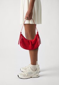 Rode netvormige handtas met een gebogen vorm en een verstelbare schouderband, gecombineerd met een geplooide witte jurk en sneakers in neutrale tinten.