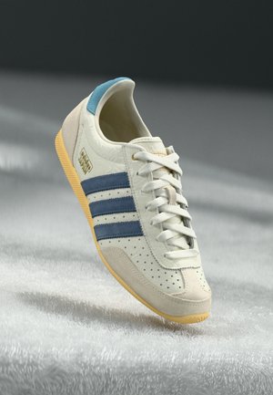 Sneaker Adidas Japon blanche avec des rayures bleues, un patch arrière bleu clair, des accents en suede beige et une semelle jaune sur une surface blanche texturée.