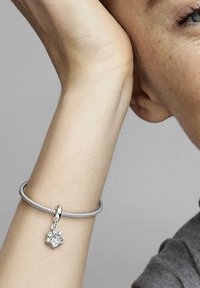 Silberne Armbanduhr mit einem verdrehten Design und einem Blumenanhänger, an dem mehrere klare Edelsteine hängen, getragen an einem Handgelenk.