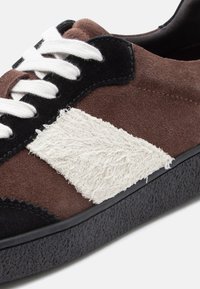 Baskets en daim avec un schéma de couleur marron et noir, présentant un accent latéral texturé blanc, des lacets blancs et une semelle en caoutchouc noire.