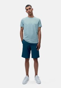 Gaiši zils kokvilnas T-krekls ar smalku dizainu, apvienots ar tumši zaļiem šortiem. Modelis valkā baltos sporta apavus, rokas atvieglinātas, stāvot uz neitrāla fona.